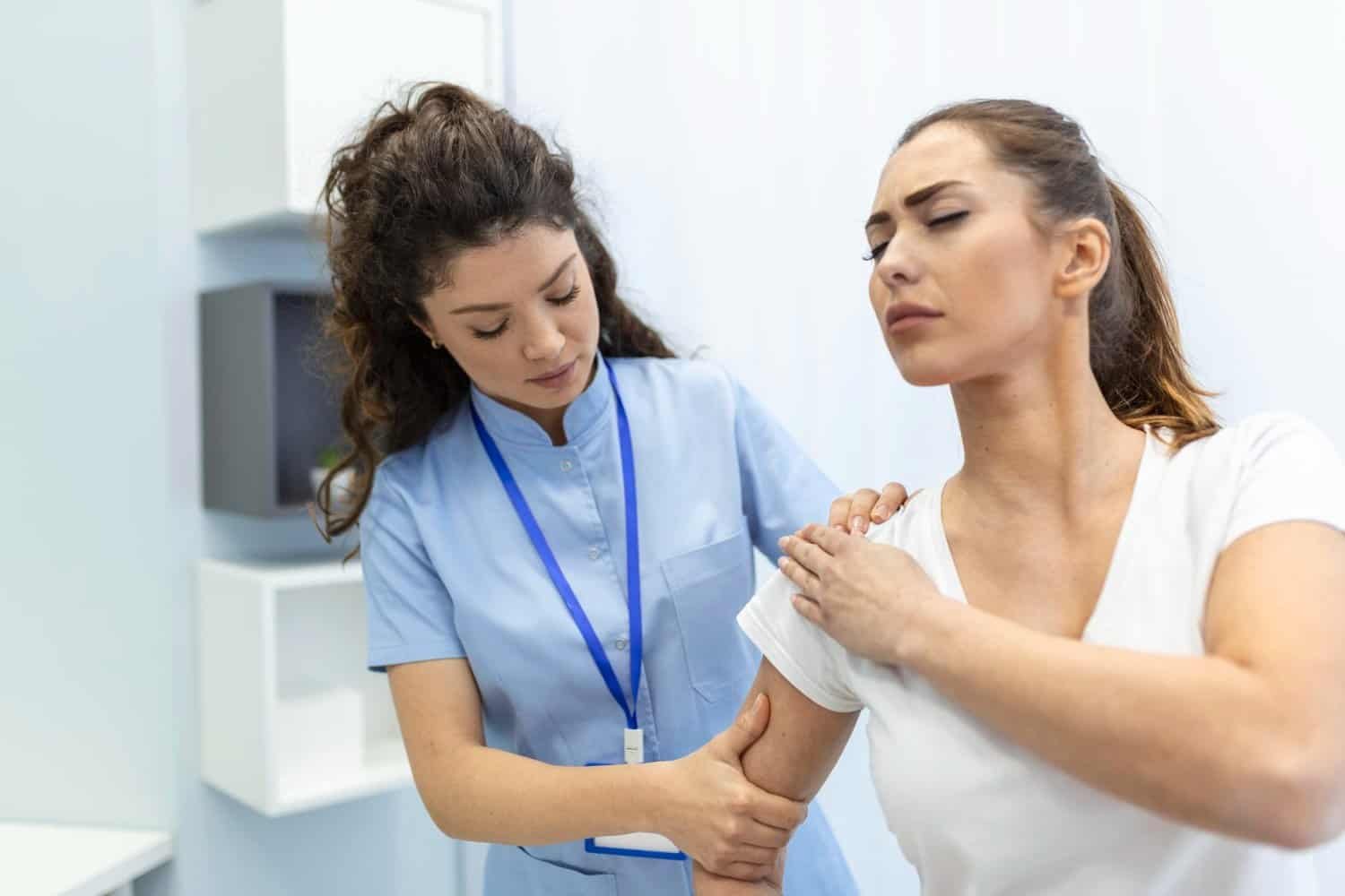 A healthcare professional examines a woman’s shoulder. The woman appears to be in pain, holding her shoulder with a pained expression while the professional gently supports and assesses her arm.