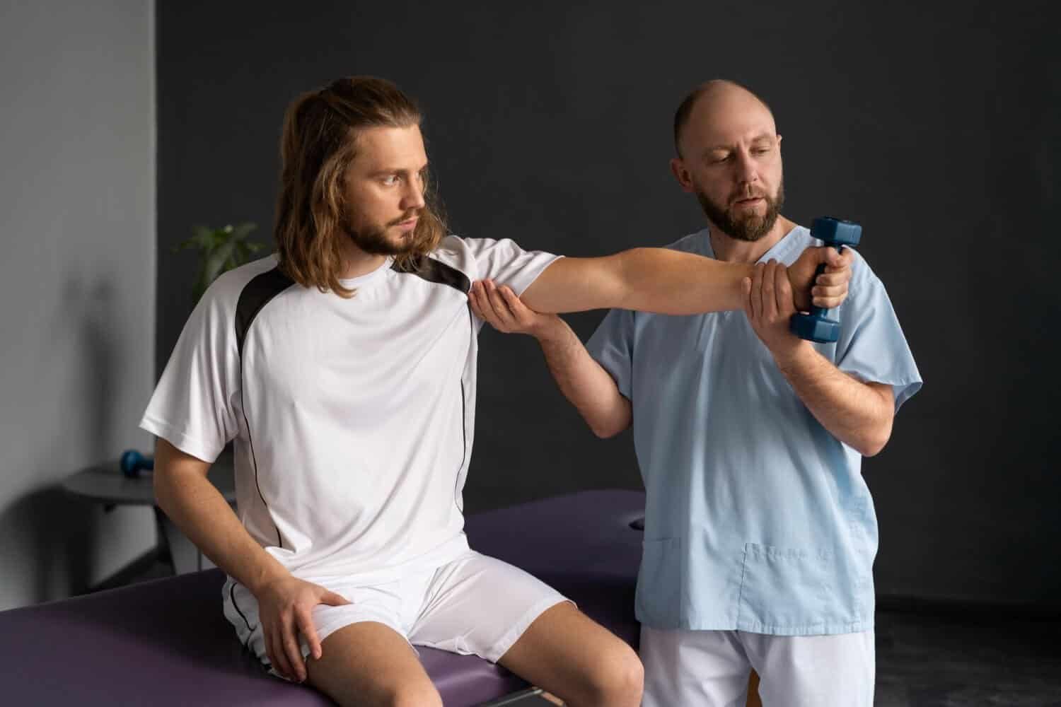 A physical therapist assists a seated man in a white sports outfit with an arm exercise using a small blue dumbbell in a clinic setting.