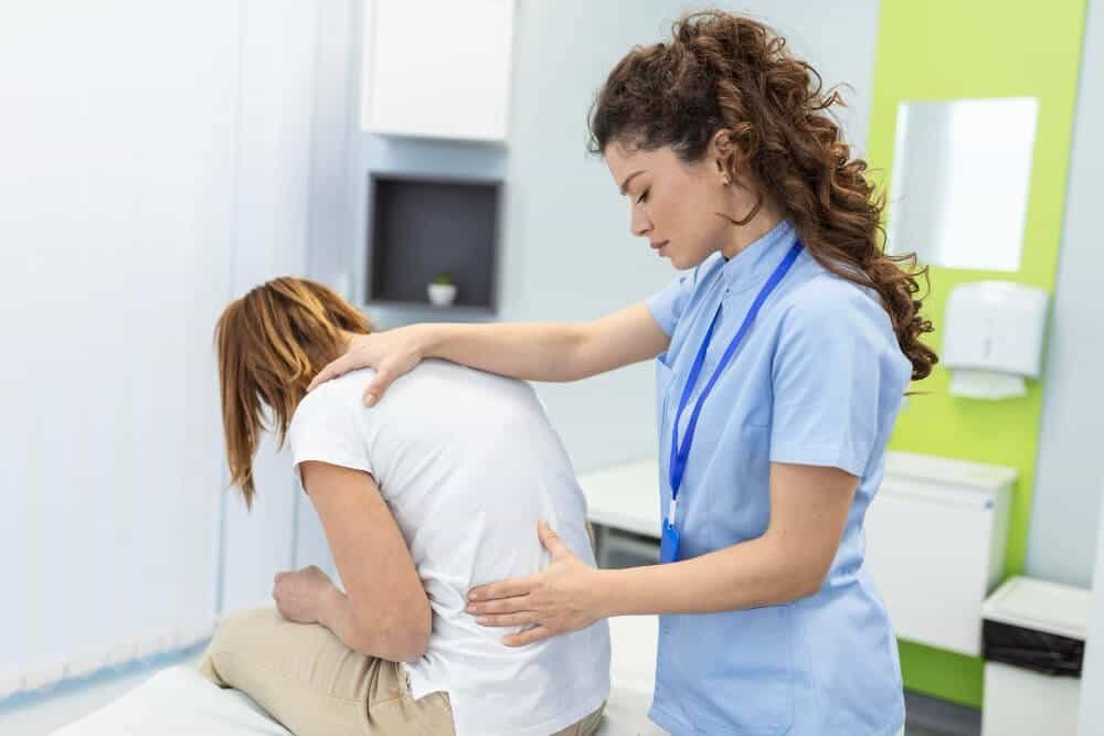 A healthcare professional examines a seated patient by gently touching their back and shoulder in a medical office. The patient appears to be experiencing discomfort.